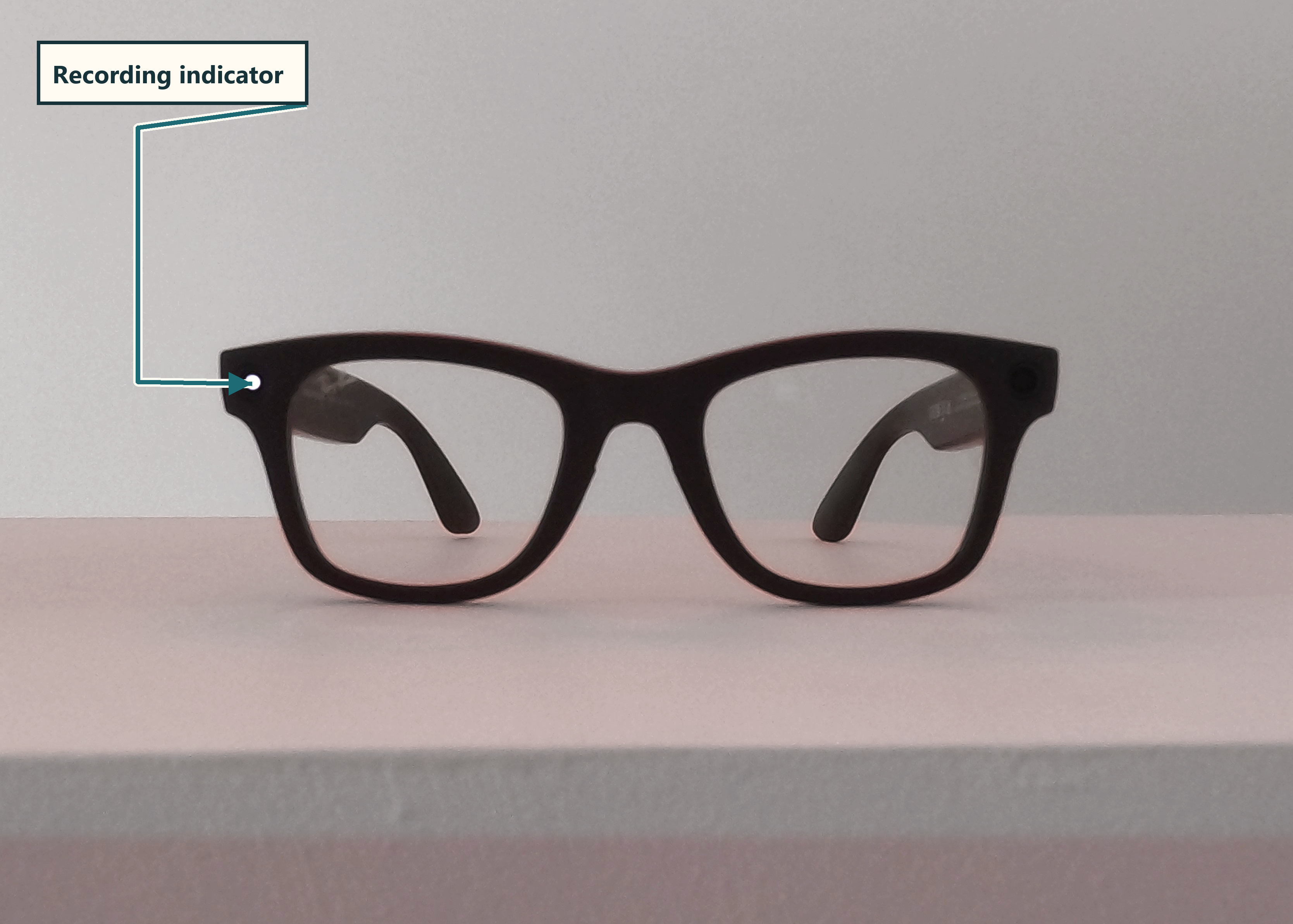 The Meta Ray-Ban smart glasses viewed from the front on a white surface. A callout arrow points to the small white recording LED on the outer corner of the left lens frame.